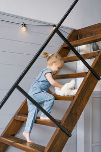 A cute two-year-old girl in a denim jumpsuit with a soft toy in her hands climbs up the stairs