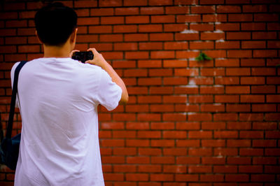 Rear view of man photographing against brick wall