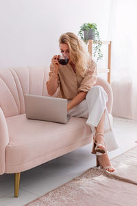 A business woman, working on laptop on sits on light pink sofa with cofee cup