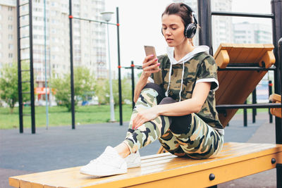 Full length of woman using mobile phone while sitting on bench at outdoor gym in city