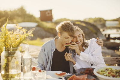 Smiling woman feeding watermelon to friend while sitting at picnic table on sunny day