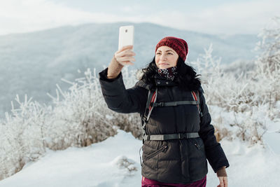 Smiling woman in warm taking selfie while standing on snowcapped mountain