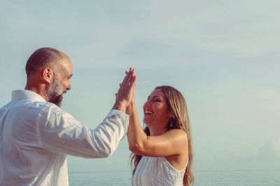 Happy couple giving high-five while standing at beach against clear sky