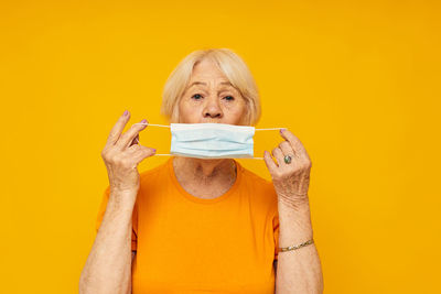 Senior woman holding surgical mask against yellow background