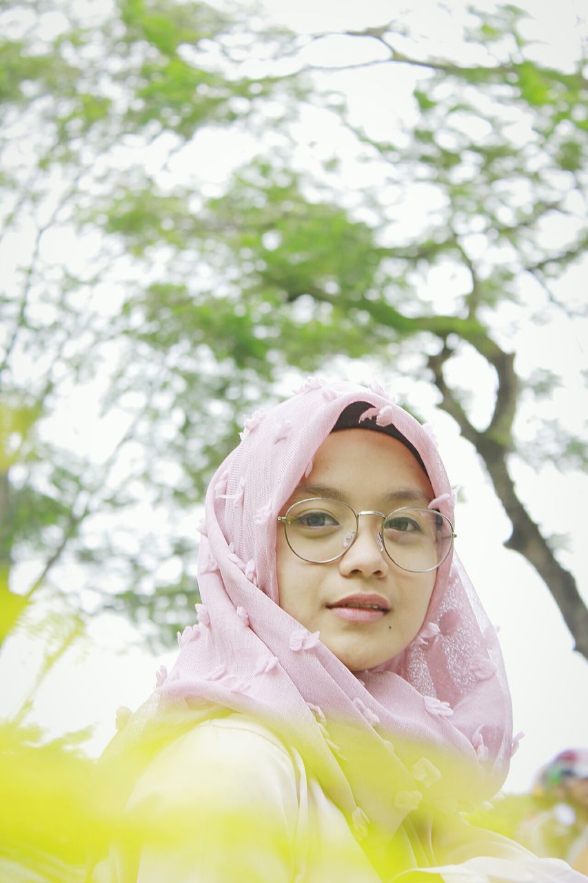 tree, real people, one person, lifestyles, leisure activity, looking at camera, day, front view, girls, focus on foreground, elementary age, outdoors, young women, young adult, portrait, eyeglasses, childhood, happiness, smiling, nature, close-up, flower