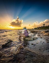 Side view of woman sitting on rock at beach during sunset