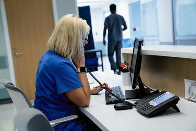 Female doctor talking on telephone while using desktop computer with male colleague in background