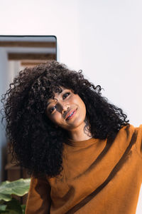 Positive young african american female with curly dark hair wearing orange sweater looking at camera against white wall