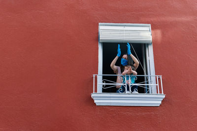 Masked mother clapping from the window with her little daughter on a red background