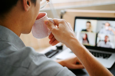 Back view of crop male entrepreneur having video conference on laptop and drinking hot beverage while discussing work issues with colleagues during coronavirus pandemic