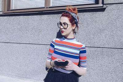 Young female hipster looking away while using mobile phone against wall