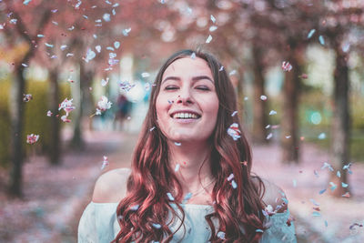 Petals falling on smiling young woman against trees