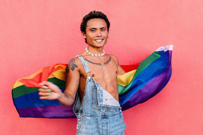 Portrait of smiling gay man holding flag against wall