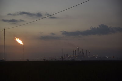 Burning flare stack and industrial buildings against sky during sunset