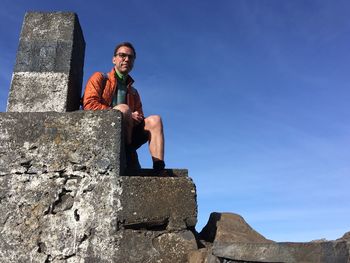 Low angle portrait of mature man standing on built structure against blue sky