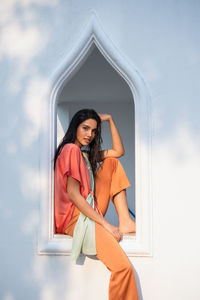Side view of cheerful charming young ethnic indian female in stylish clothes sitting on windowsill of arched window in oriental style house and looking at camera