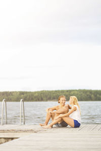 Couple sitting on boardwalk by lake against sky