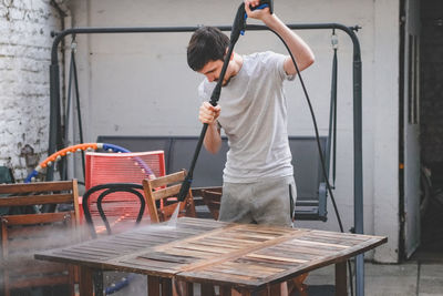 A caucasian guy in gray sweatpants and a t-shirt washes a wooden garden table with a karcher