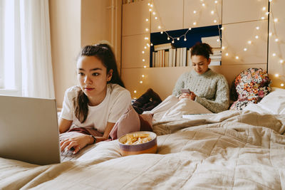 Young woman using laptop while friend using smart phone on bed at home