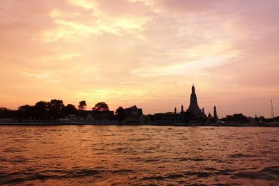 Silhouette buildings by river against sky during sunset