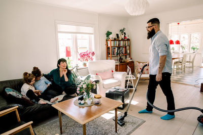 Man looking at family while cleaning carpet with vacuum cleaner in living room