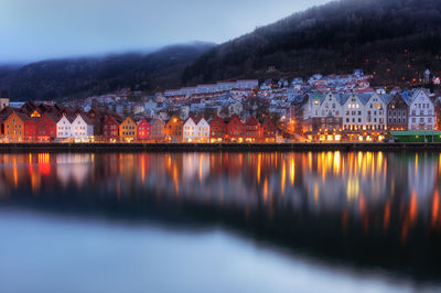 Illuminated buildings by lake against sky at dusk