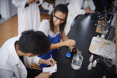 High angle view of smiling multi-ethnic students mixing solutions and writing in spiral notepad at chemistry laboratory