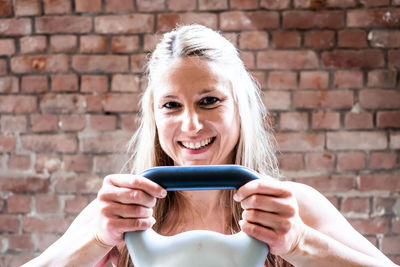 Portrait of smiling woman holding kettle bell against brick wall