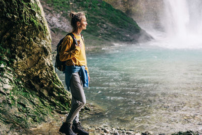 Active female tourist with a backpack in outfit in a mountainous area near a stream and a waterfall.