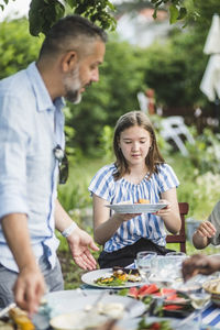 Girl holding food plate while sitting at table with family in backyard