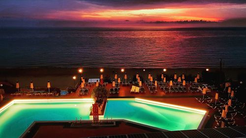 Illuminated swimming pool by sea against sky during sunset