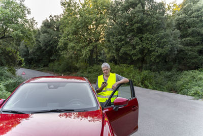 Senior man standing at his broken car using his smartphone