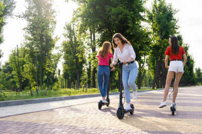 Three young girl friends on vacation having fun driving electric scooter through the city park 