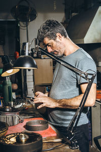 Middle aged male silversmith shooting metal ring via cellphone while standing near workbench during work in professional workshop