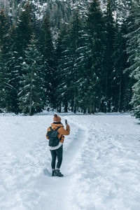 Rear view of female hiker standing on snowy path, taking photos of moody forest