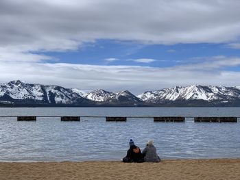 People sitting on snowcapped mountain by lake against sky