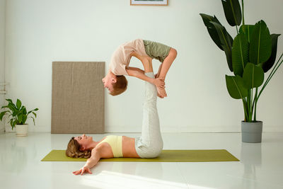 A woman, doing yoga with a child boy, lies on a sports mat, raising her legs up