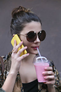 Portrait of young woman drinking smoothie while telephoning with smartphone