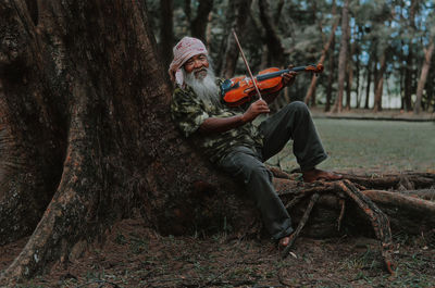 Portrait of man playing violin while sitting against tree trunk