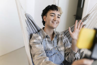 Positive young female with dyed hair waving hand while shooting live video on smartphone in hammock in light room