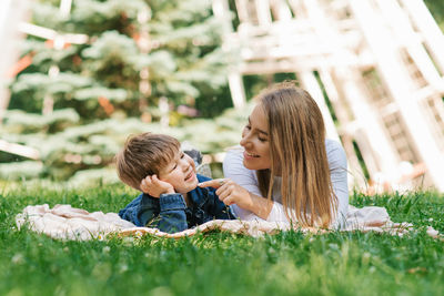 Young caucasian mother and her young son lie on a blanket on the grass in the park in summer