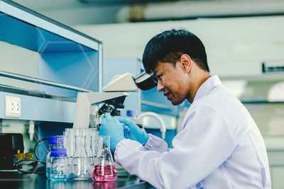 Asian man lab technician in protective glasses and gloves sits next to a microscope in laboratory.