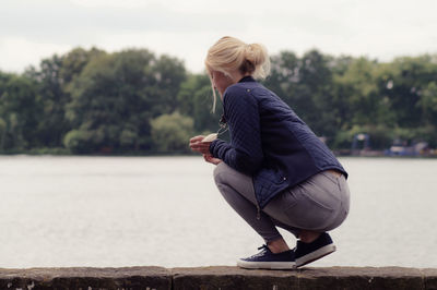 Beautiful woman crouching on retaining wall by river against sky