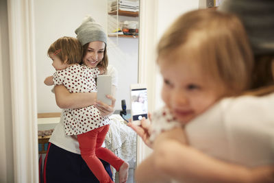 Smiling podcaster taking selfie with daughter through smart phone while reflecting on mirror