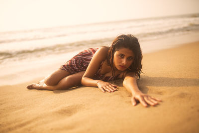 Portrait of young woman lying on sand at beach against sky