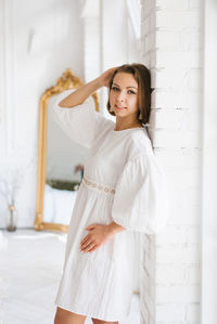 Young woman in a white dress stands near a white wall in the house