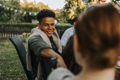 Smiling young man sitting on chair while talking with female friend in back yard at dinner party