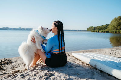 Snow-white japanese spitz kissing woman with locs, female dog owner sitting on beach of city lake