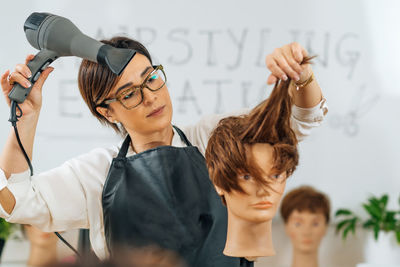 Hairstylist with the hair dryer in hands explaining hairstyling techniques, using mannequin head