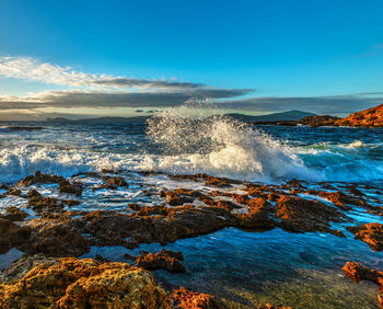 Waves splashing on rocks at shore against blue sky